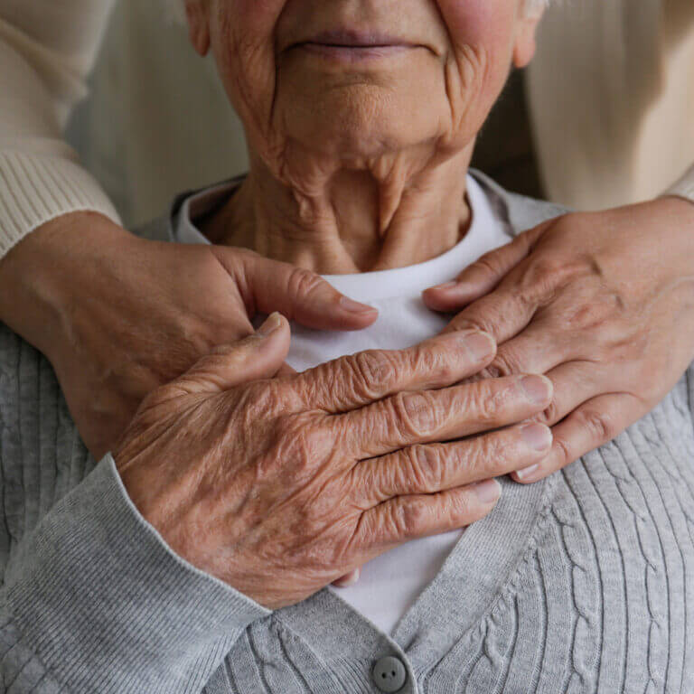 An older woman holding her hands to her chest, with a carer’s hands on her shoulders
